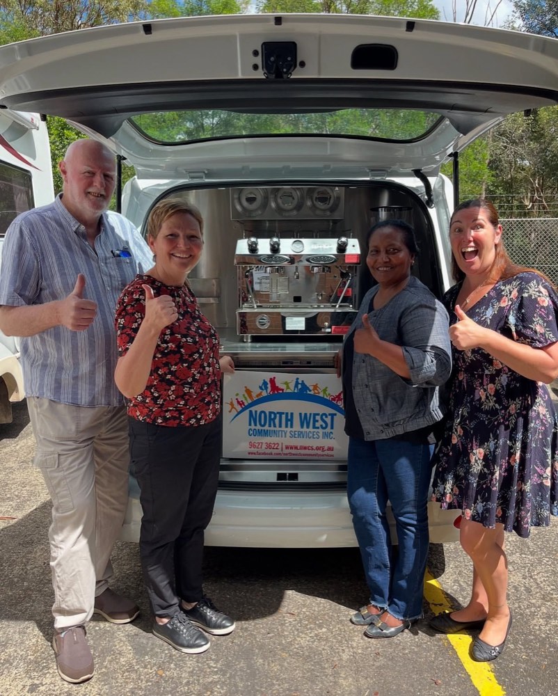Four people giving a thumbs up at the open rear hatch of a white coffee van, a two-group espresso machine on the bench behind them and a North West Community Services sign across the counter front.