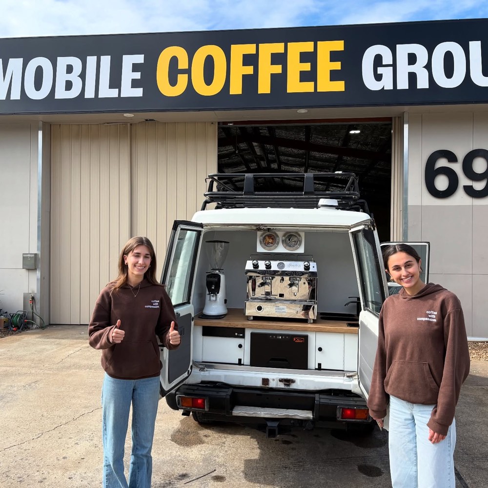 Two people in brown hoodies giving a thumbs up beside a white LandCruiser ute fitted out with an espresso machine and grinder, parked under The Mobile Coffee Group workshop sign.