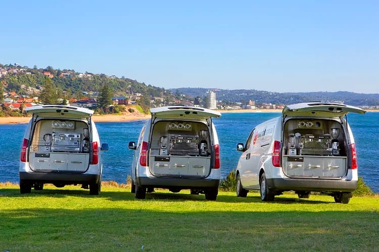 Three white coffee vans parked on grass above a beach, rear hatches raised over their stainless benches.