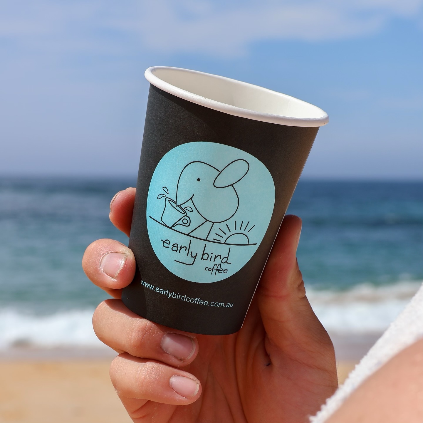An Earlybird cup held against a blue sky at the beach.