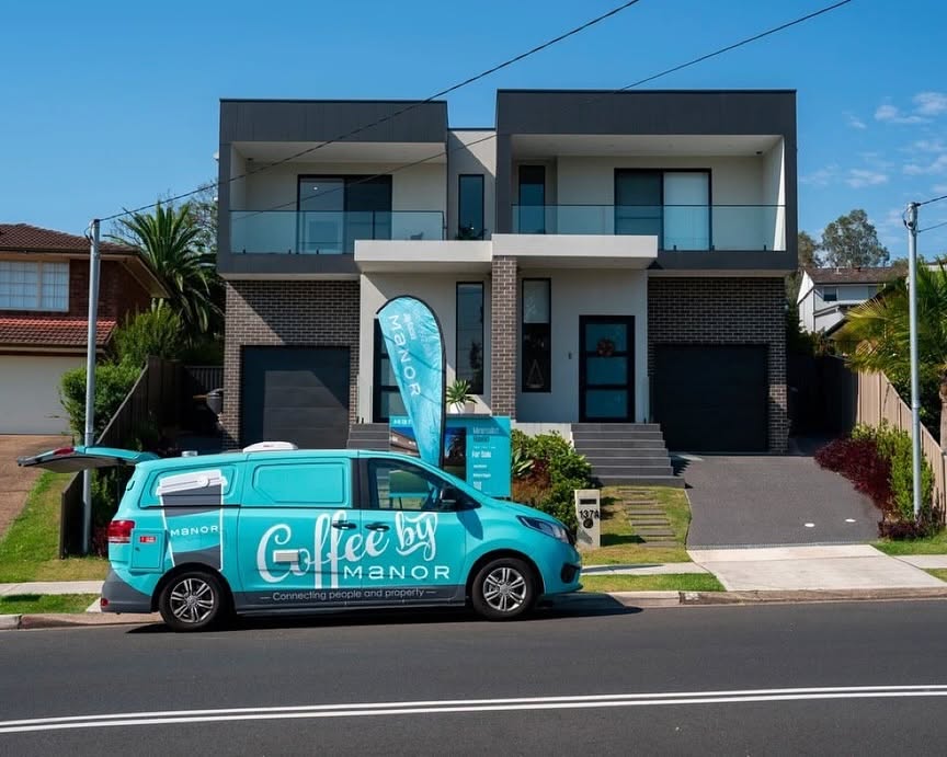 A teal Coffee by Manor van parked outside a new two-storey house beside a for-sale flag.