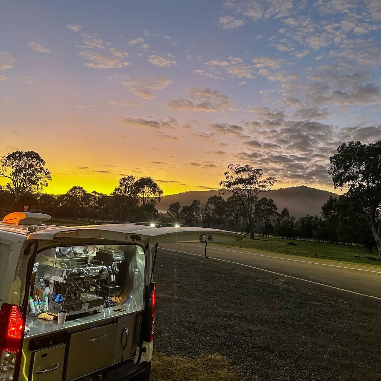 A coffee van open at the roadside in farmland, its lights on under a yellow and violet dawn sky.