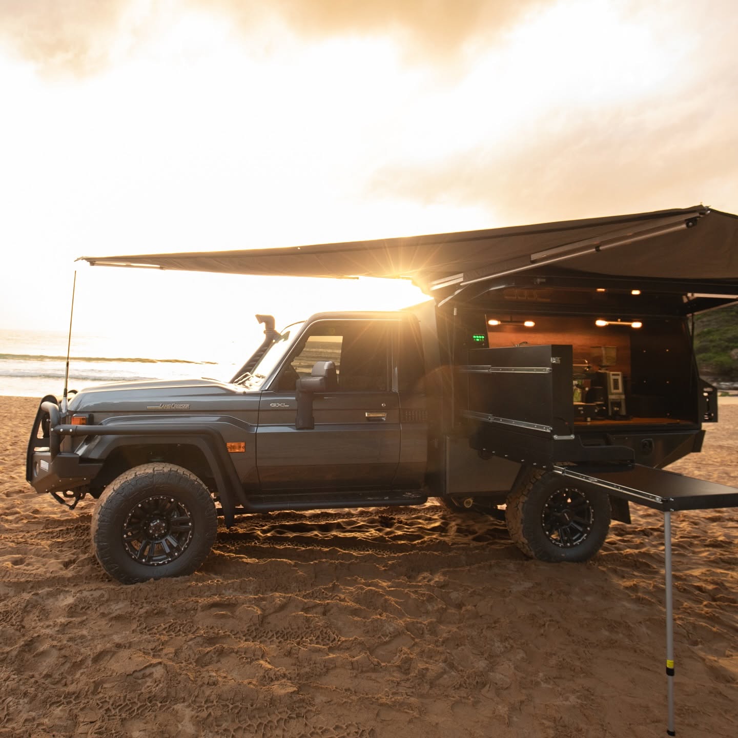 A black LandCruiser ute with its awning out and canopy open on a beach at sunrise, the sun flaring behind it.