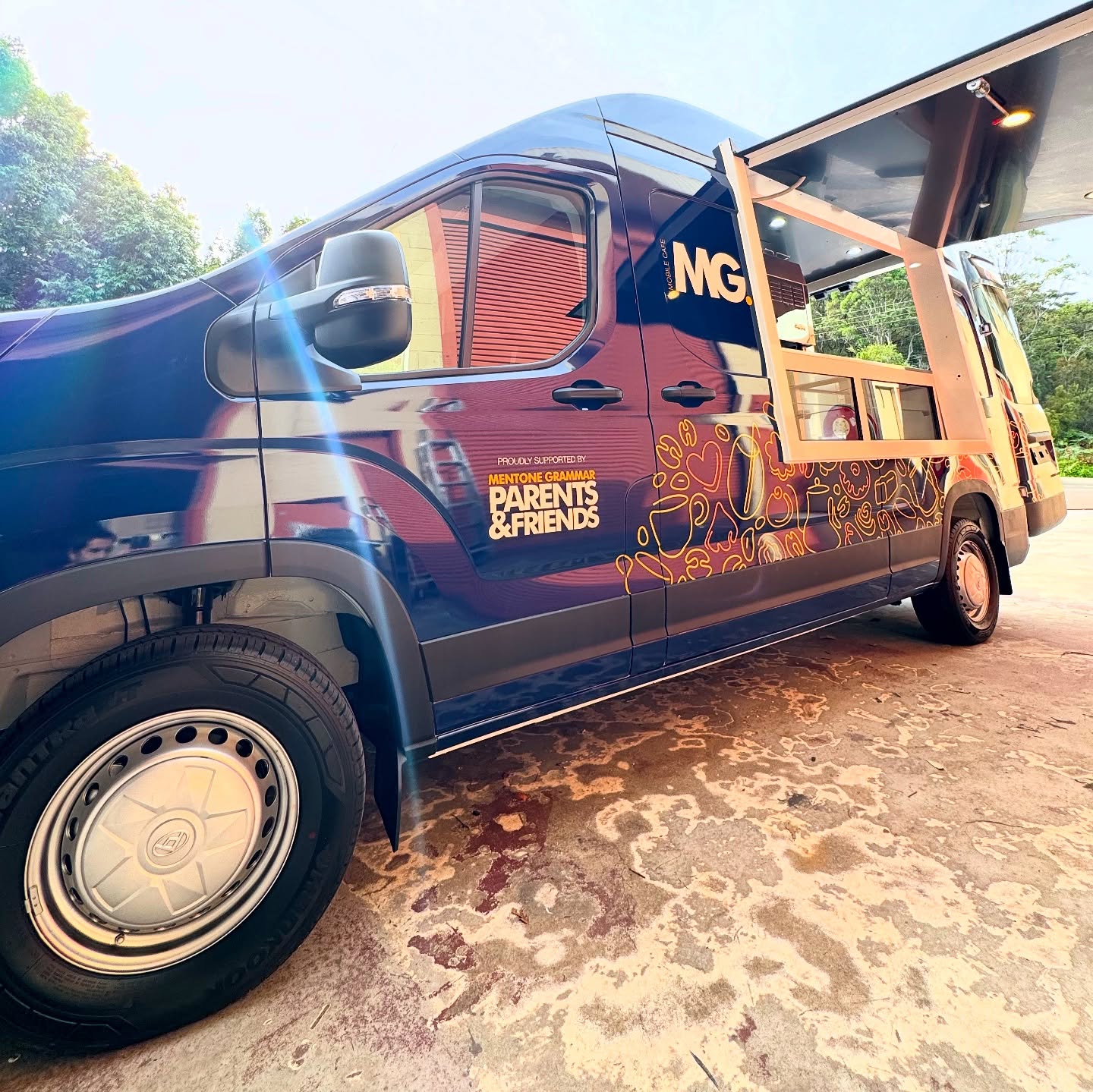 A navy mobile cafe van with a timber servery frame folded out, parked on a gravel drive.