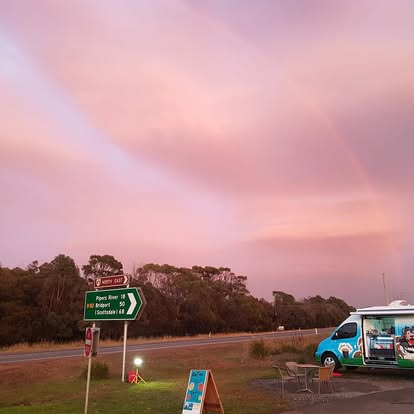 A coffee van set up at a country highway junction under a rainbow and a pink dusk sky.