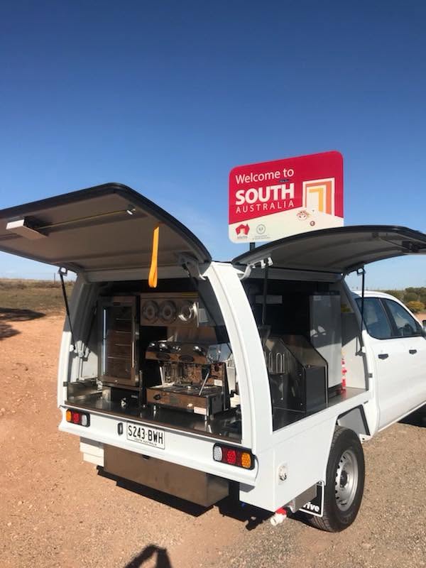 A white coffee ute open at the roadside beneath the Welcome to South Australia border sign.