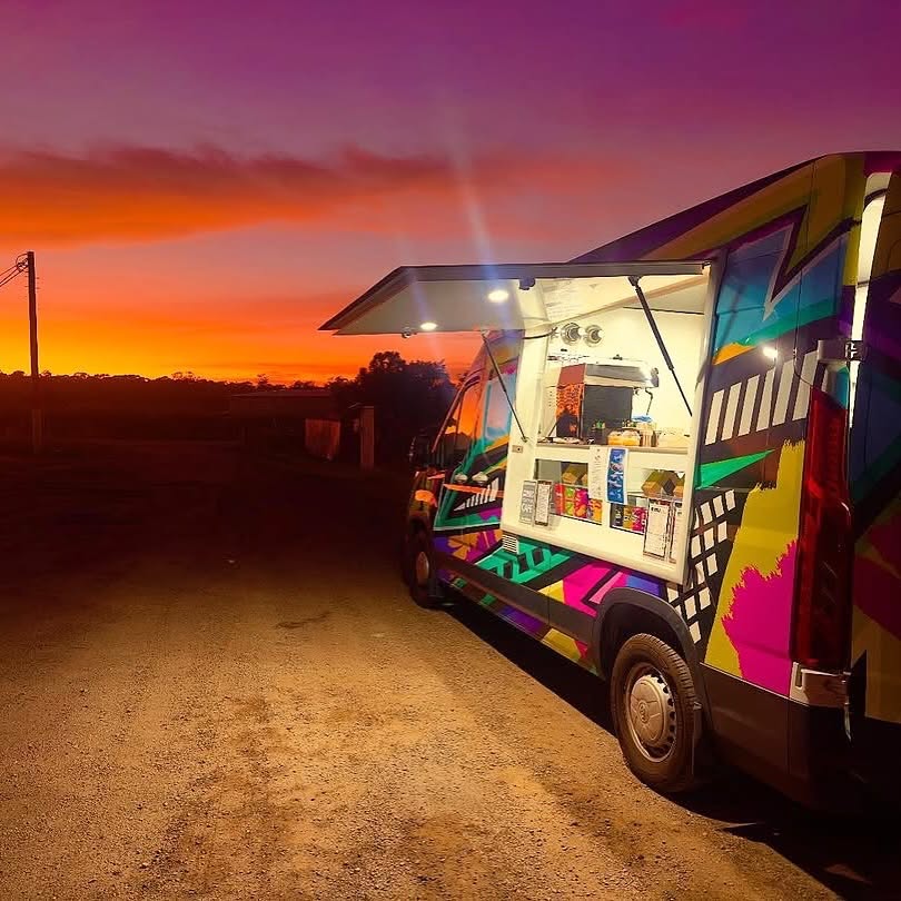 A brightly wrapped coffee van trading from a gravel roadside against an orange and purple sunset.