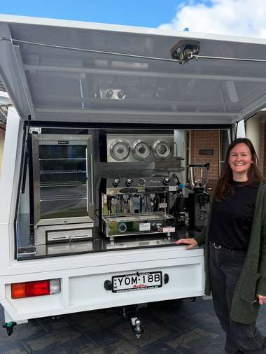 An operator standing at the open rear of her white coffee van, the bench and grinders visible inside.