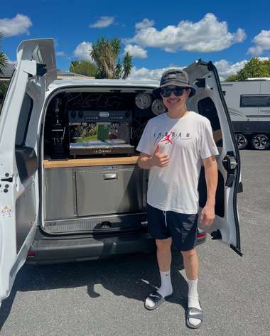A young operator in a bucket hat holding a takeaway cup beside his newly fitted coffee van.