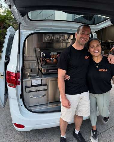 Two operators standing arm in arm at the rear of their white coffee van.