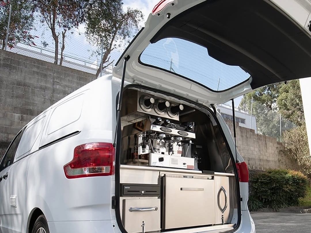 Close view of a rear-hatch fit-out: espresso machine and grinder on the bench, cup warmers set into the ceiling panel, and cabinetry with a fridge drawer beneath.