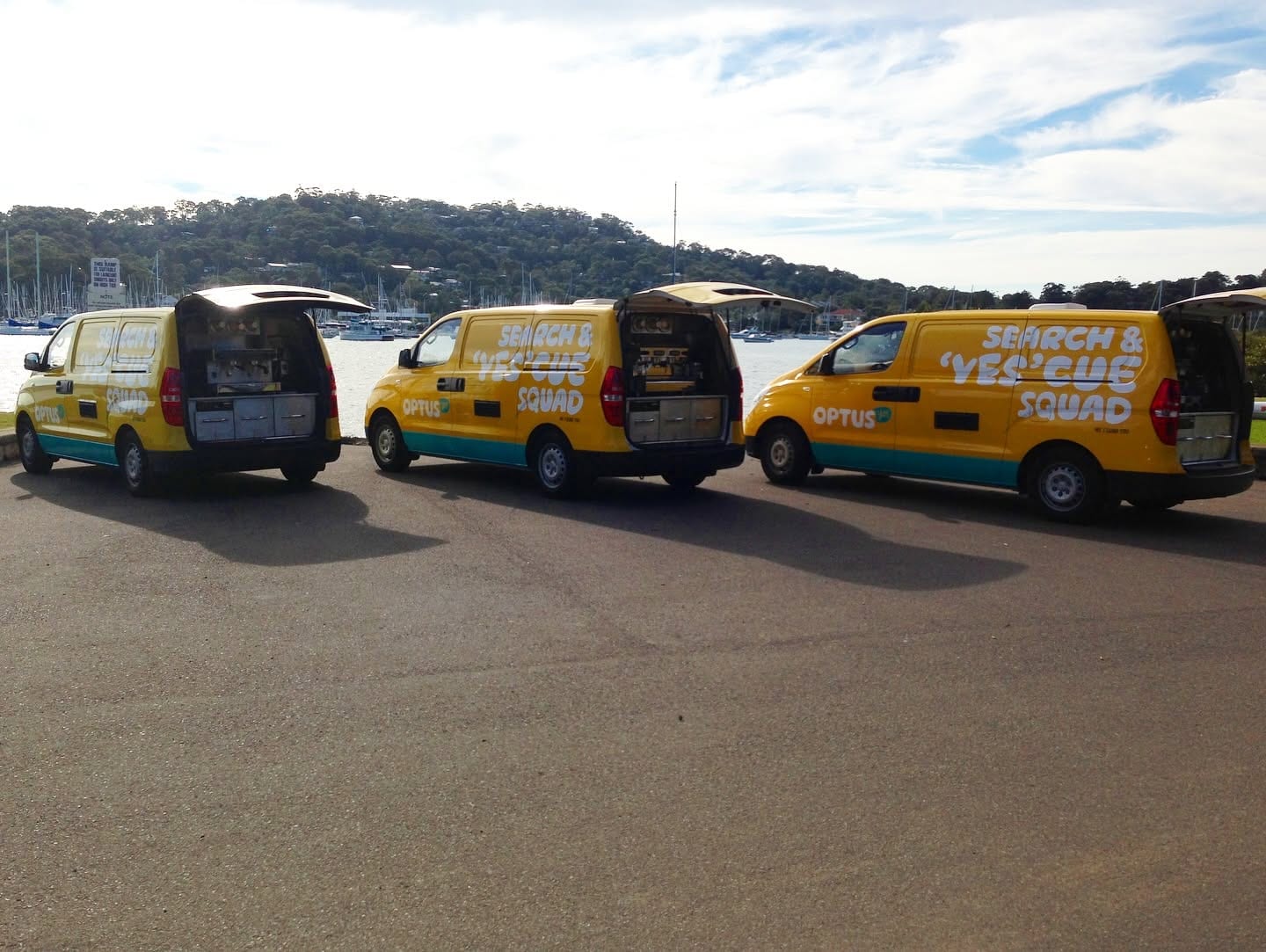 Three yellow Optus-liveried vans parked in a row beside the water, rear hatches raised over their coffee benches.