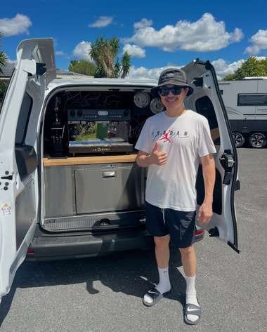 A man in a bucket hat gives a thumbs up beside a white van, its rear hatch raised over an espresso machine and stainless bench, caravans parked behind.