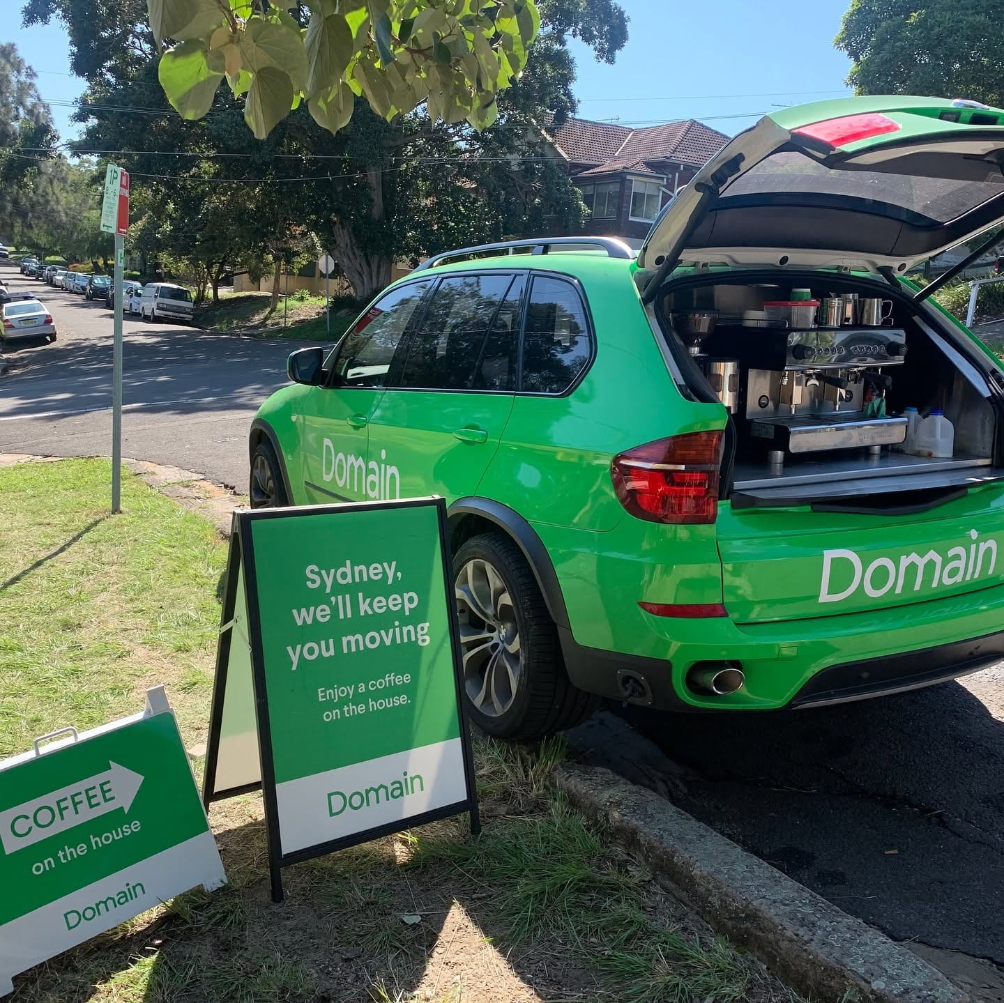 A bright green Domain-branded BMW X5 with the boot open on a suburban verge, A-frame signs offering coffee on the house.