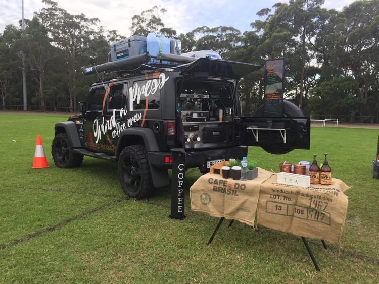 A black Jeep set up on a sports field with an awning out, espresso machine in the back and a trestle table of coffee gear.