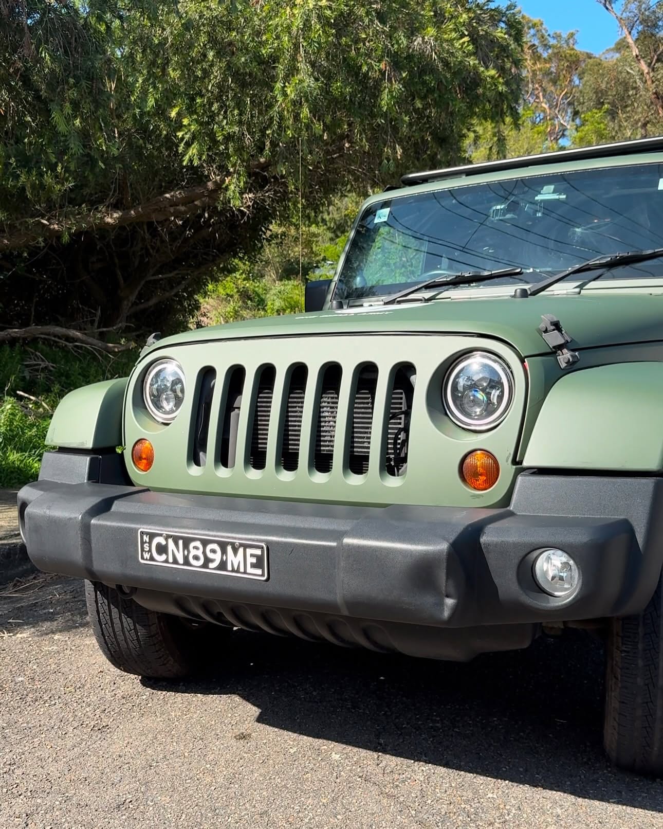 A matte olive-green Jeep Wrangler parked on gravel under trees, seen head-on.