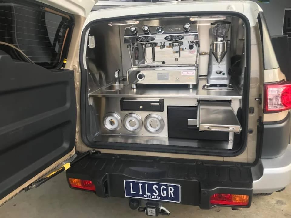 A gold Mitsubishi Pajero with its rear hatch raised over a stainless bench, two-group espresso machine, grinder and recessed cup wells.